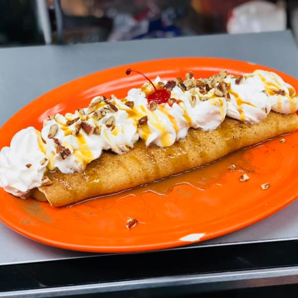 A long, golden crepe topped with whipped cream, caramel drizzle, pecans, and a cherry on an orange plate.